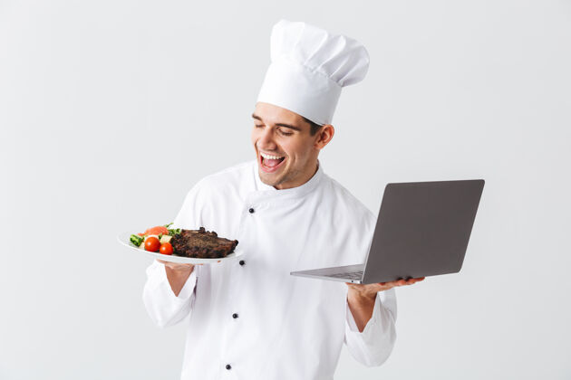 便携式兴奋的厨师穿着制服站在白色的墙上 拿着笔记本电脑 展示着一道菜服务老板盘子