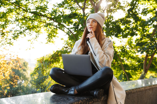 坐着一个穿着秋衣 戴着秋帽 坐在户外 用笔记本电脑沉思的年轻女子女人小玩意目标