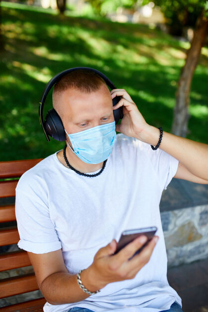 互联网男子戴着耳机听音乐 在公园户外用智能手机戴着护面面具 生活方式新常态疾病病毒2019ncov