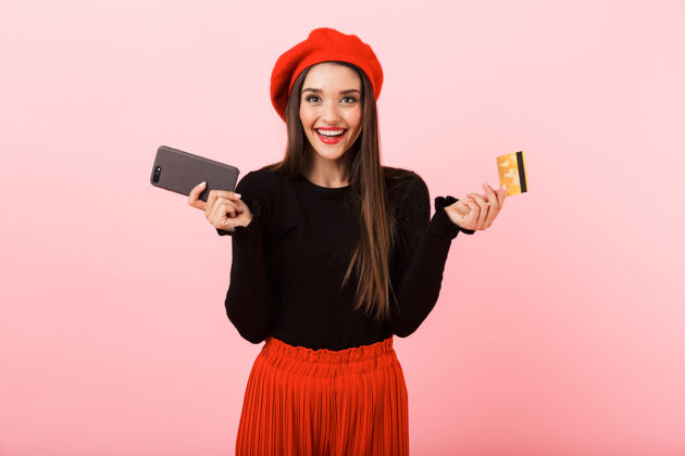 女性一个戴着红色贝雷帽 手持手机和塑料信用卡 孤立地站在粉色背景下的快乐美丽的年轻女子的画像信用年轻消费主义
