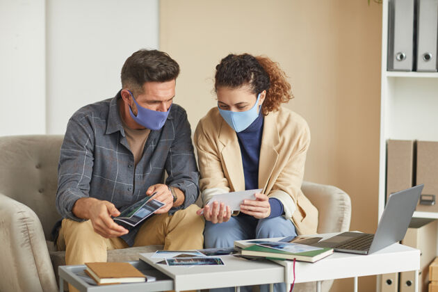 计算机两个戴着面具的设计师坐在沙发上 在办公室的团队合作中检查照片讨论坐着团队合作