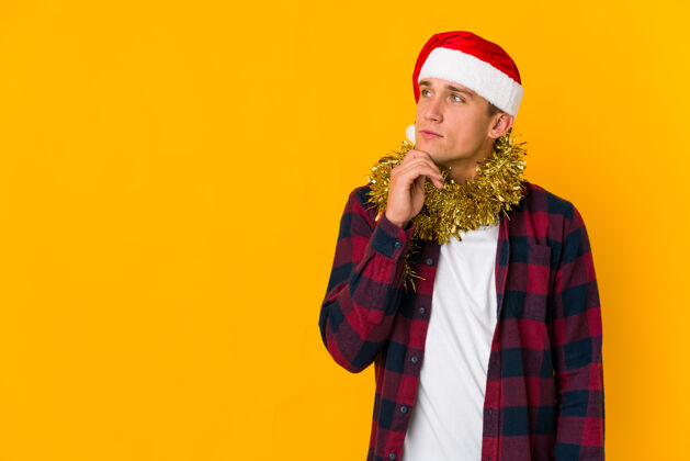 男人戴着圣诞帽的白人年轻人拿着一件礼物 孤立地站在黄色背景上 带着怀疑和怀疑的表情侧视着不确定思想十二月