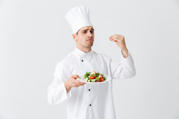 膳食严肃的男厨师穿着制服 在白色墙壁上的盘子上展示新鲜的蔬菜沙拉专业烹饪盘子