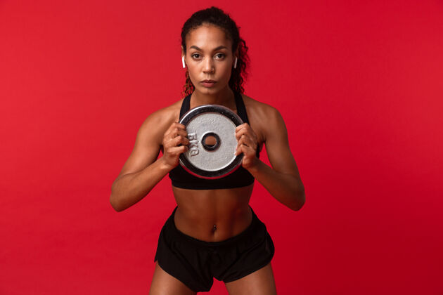 温暖身着黑色运动服的非裔美国女运动员手持杠铃 隔着红墙教练体操运动