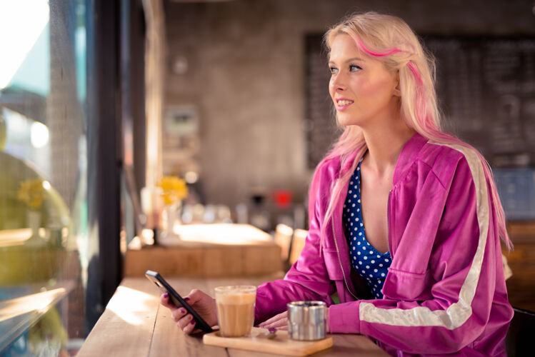 优雅快乐的年轻漂亮的金发女人在咖啡店用手机思考科技信息人