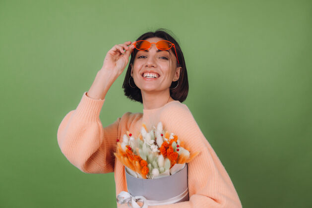 积极年轻女子穿着休闲的桃色毛衣隔离在绿橄榄墙上捧着橘白色的花盒组成的棉花 团花 小麦和紫罗兰为礼物开心惊喜年轻时尚室内