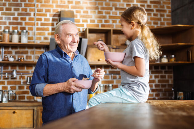 室内积极的老人一边吃谷类食品一边享受和孙女在一起的时光成人桌子家庭