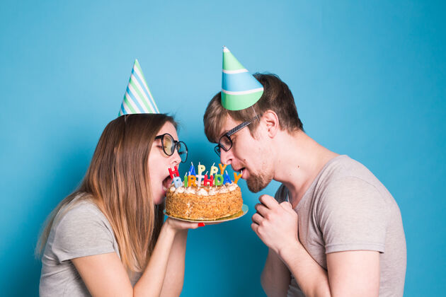 搞笑一对奇怪的年轻夫妇戴着纸帽子的女孩和男孩想咬下一块祝贺蛋糕表情明亮甜点