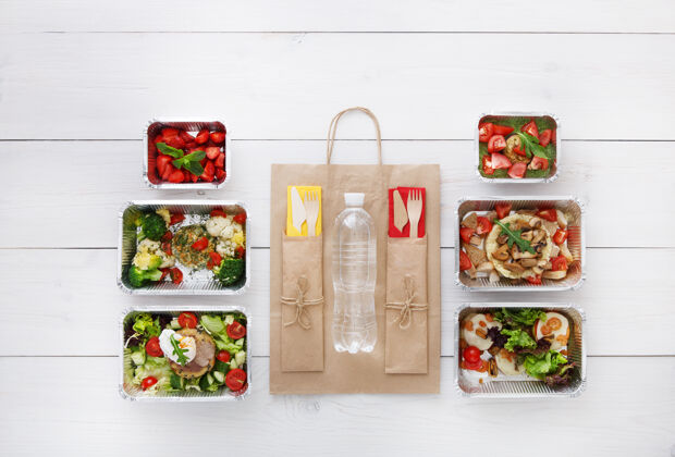 早餐健康食品配送外卖食品铝箔盒中的蔬菜 肉类和水果 餐具 水和牛皮纸包装俯视图 平放在白色木头上 有复印空间盒子容器自然