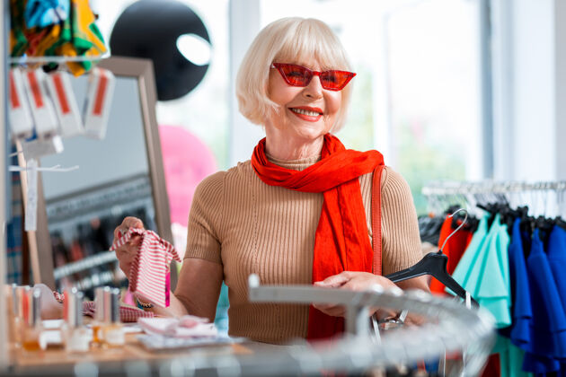 老年人很高兴与商店收集照片中的快乐的老妇人透过红色太阳镜看着别处 同时选择一个头带来搭配她的衣服欢呼衣服心情