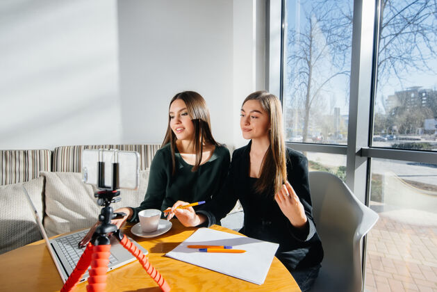年轻人两个年轻漂亮的女孩坐在咖啡馆里 录制视频博客 在社交网络上交流数码女商人讨论
