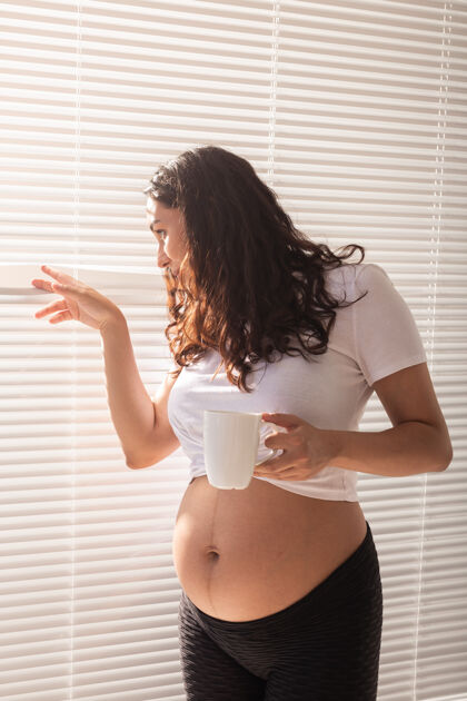 产妇好奇的年轻漂亮的孕妇喝着茶 透过百叶窗望着窗外肚子中年放松