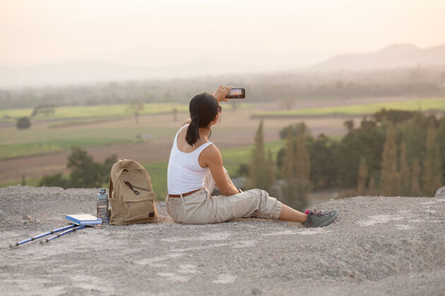 黑年轻女子拿着手机坐着夕阳下的高山旅游小道活动移动白