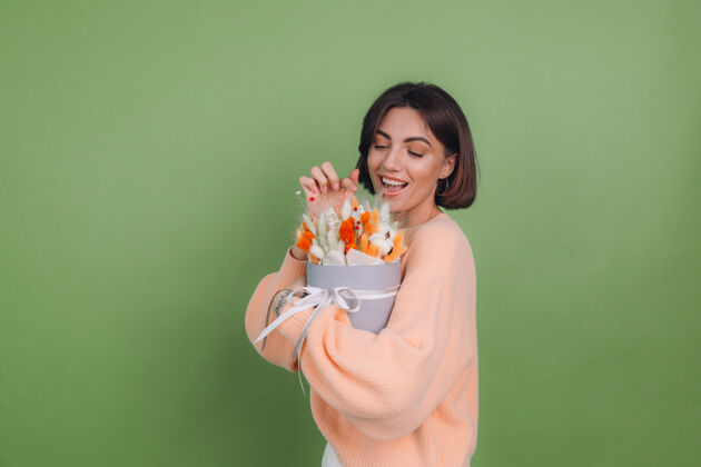 女人年轻女子穿着休闲桃色毛衣隔离在绿橄榄墙上手持橙白色花盒组成的棉花 吉普赛拉小麦和拉古鲁斯作为礼物开心惊喜时尚年轻时尚