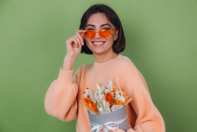 时尚年轻女子穿着休闲的桃色毛衣隔离在绿橄榄墙上捧着橘白色的花盒组成的棉花 团花 小麦和紫罗兰为礼物开心惊喜快乐魅力开花