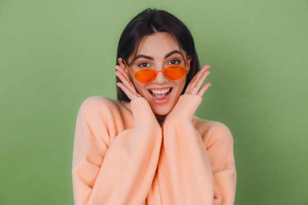 天哪穿着休闲桃色和橙色眼镜毛衣的年轻女子隔离在绿橄榄墙上兴奋地张嘴摊开双手成功人兴奋