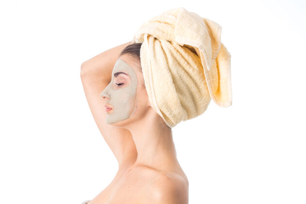 美丽年轻可爱的女人用洁面面膜和毛巾在白色的墙上隔离在头上护理她的皮肤护理面部服用