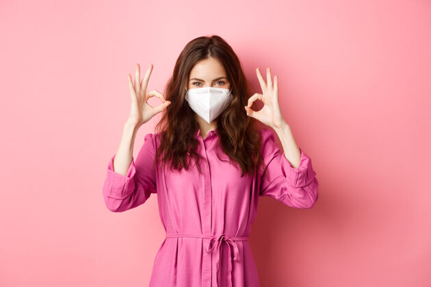 女性Covid-19 流行性疾病和生活方式的概念开朗的女孩表现出良好的迹象 戴医用呼吸器作为预防电晕的措施 粉红色的墙壁乐观社交距离快乐