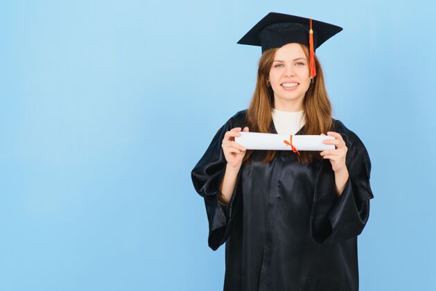喜悦戴着毕业帽和毕业礼服的女研究生 背景是蓝色的学士斗篷学习