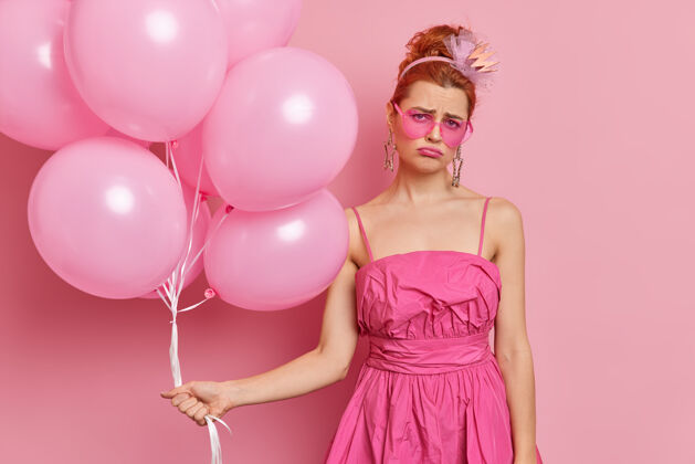 不高兴不满意的红发女人看起来很悲伤穿着喜庆的裙子拿着一堆气球在生日派对上感到不开心和孤独隔着粉色的墙孤独地为变老而悲伤太阳镜年轻不高兴