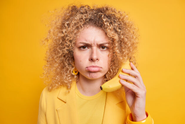 愤怒特写镜头：闷闷不乐的卷发女人拿着香蕉 电话放在耳边假装打电话给某人 表情不悦 穿着黄色衣服 在室内摆出负面情绪的姿势水果不满不高兴