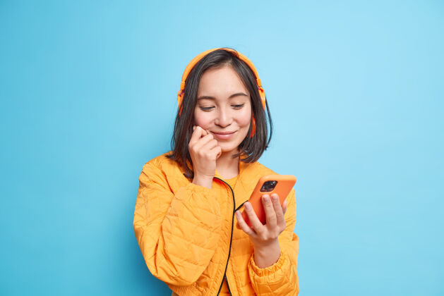 漂亮心满意足的亚洲女孩 深色头发 专注于智能手机屏幕 从播放列表中选择音频曲目 戴着无线立体声耳机 戴着橙色夹克 隔着蓝色的墙壁高兴使用手机