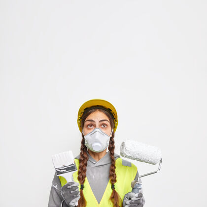 头盔建筑和施工概念惊讶的女人扎着两条辫子 戴着防护面具和安全帽 穿着制服 建筑设备集中在隔离的白墙上土木建造师刷子