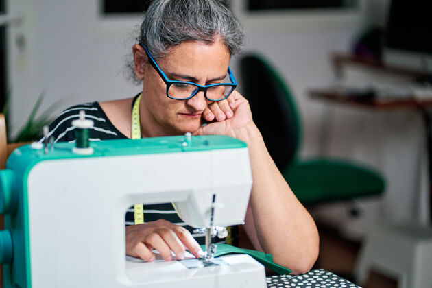 工作沉思的白发中年妇女在家里用缝纫机缝纫线车间护目镜