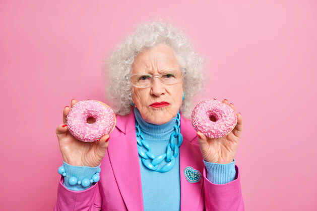 表情不满满脸皱纹的老太太拿着两个美味的甜甜圈吃垃圾食品穿时髦的衣服粉红时尚皱眉