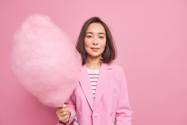亚洲漂亮的亚洲女人拿着加糖的粉色棉花糖 穿着正式的衣服和孩子们一起度假 吃高热量的甜点 隔着玫瑰色的墙壁生活方式的概念高兴享受肖像