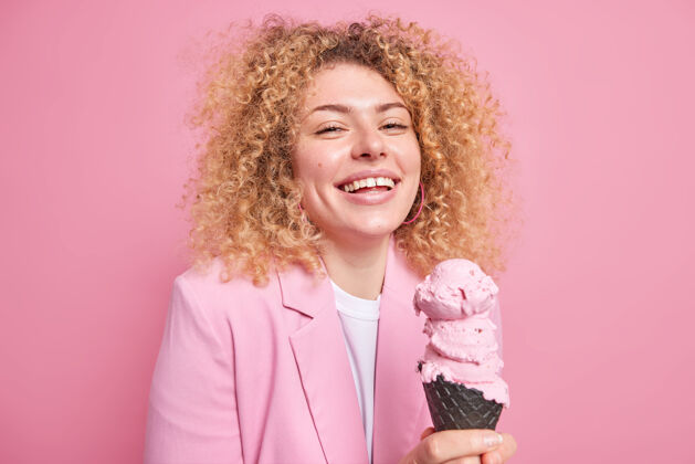 味道快乐漂亮的卷发女人喜欢吃美味的草莓冰淇淋有愉快的表情无忧无虑的生活方式表达真实的情感穿着优雅隔离在粉红色的墙上食物美味高兴