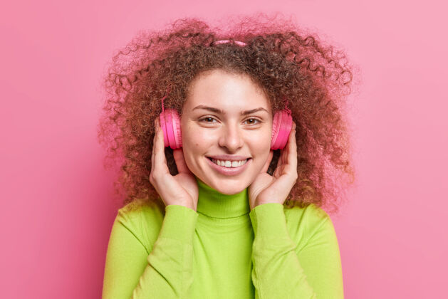 快乐帅气的欧洲女人meloman的头像卷曲的头发戴着立体声耳机听音轨有着乐观的心情穿着休闲的高领毛衣隔着粉色的墙壁旋律积极欢呼