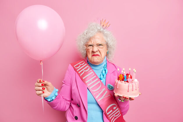 情绪不满意的卷发高级女士庆祝生日穿着时尚的衣服和珠宝摆出充气气球美味的蛋糕看起来脾气暴躁退休人物不满意