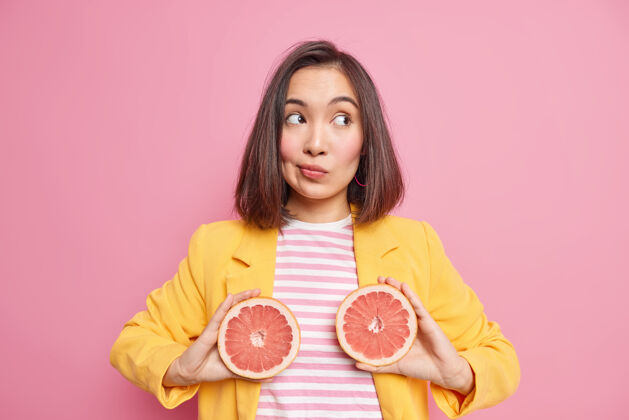 一半体贴的亚洲女人的水平镜头拿着两个葡萄柚半集中在一起穿着时尚的衣服隔离在粉红色的墙上天然营养和多汁的柑橘水果概念黑发自然漂亮