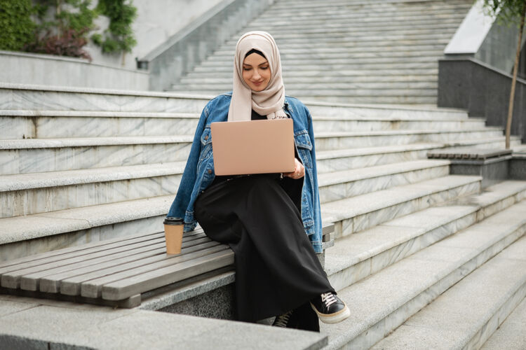 伊斯兰现代时尚的妇女戴着头巾 穿着牛仔夹克和黑色长袍坐在城市街道上用笔记本电脑工作职业头巾