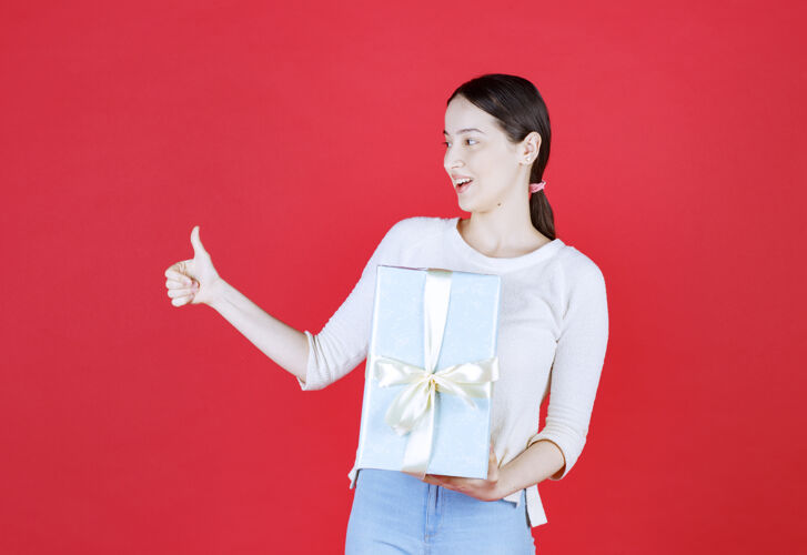 手势震惊的年轻女子拿着礼品盒 并示意拇指向上模特快乐包装
