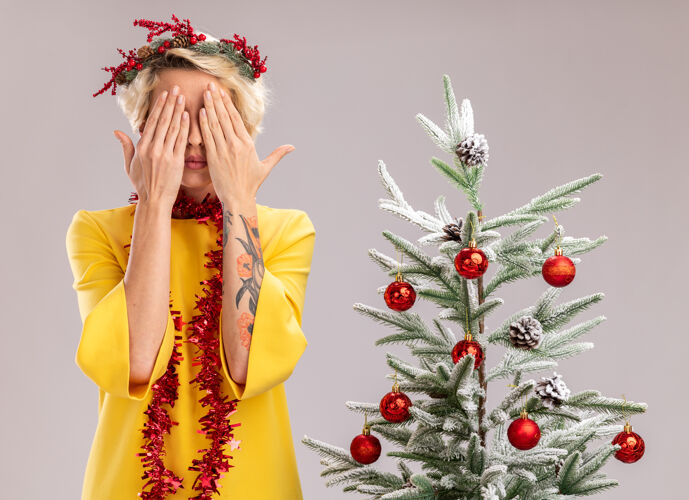 年轻年轻的金发女人戴着圣诞花环 脖子上戴着金属丝花环 站在装饰好的圣诞树旁 双手孤立地站在白色的墙上 遮住眼睛脖子金箔圣诞节
