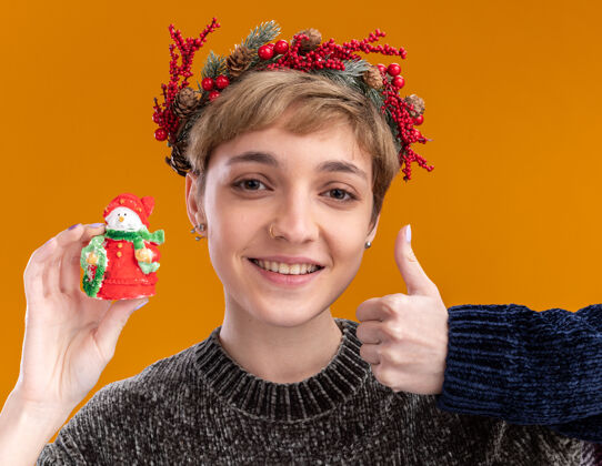 美丽微笑着的年轻漂亮女孩戴着圣诞花环 手持小圣诞雪人雕像 竖起大拇指 孤零零地站在橙色的墙上雪人微笑雕像