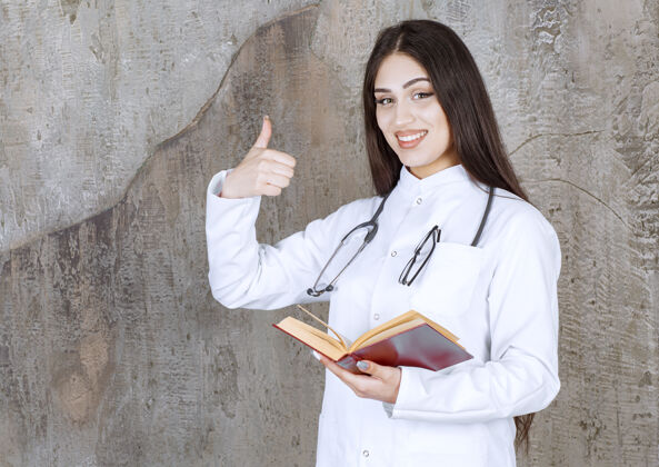 外套年轻的女医生在灰色的墙上竖起大拇指严重女性医院