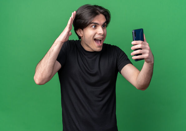 感觉兴奋的年轻帅哥穿着黑色t恤拿着手机 看着隔离在绿色墙上的手机电话脸人