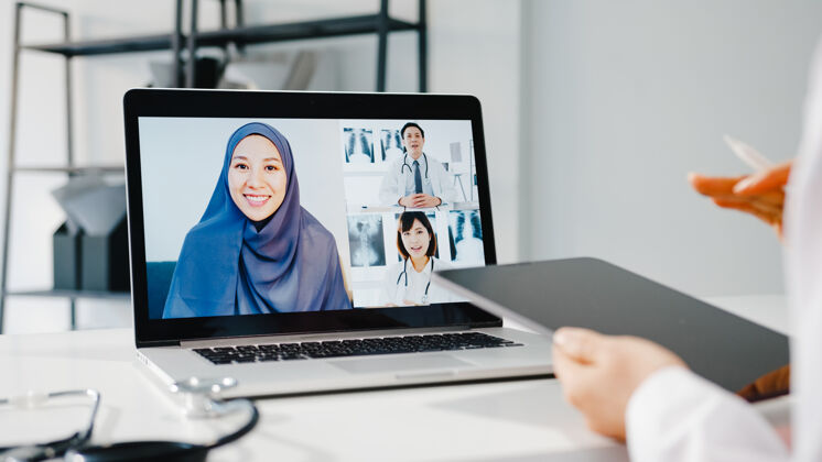 头巾年轻的亚洲女医生穿着白色医疗制服 手持听诊器 使用笔记本电脑进行视频电话会议电脑团队网络研讨会