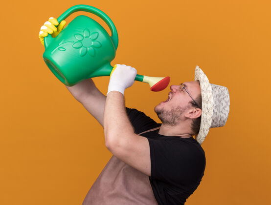 站快乐的年轻男园丁戴着园艺帽和手套拿着浇水罐在橙色的墙上给自己浇水衣服姿势感情