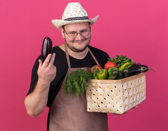 花园高兴的年轻男园丁戴着园艺帽 手里拿着蔬菜篮子 把茄子隔离在粉红色的墙上男年轻人茄子