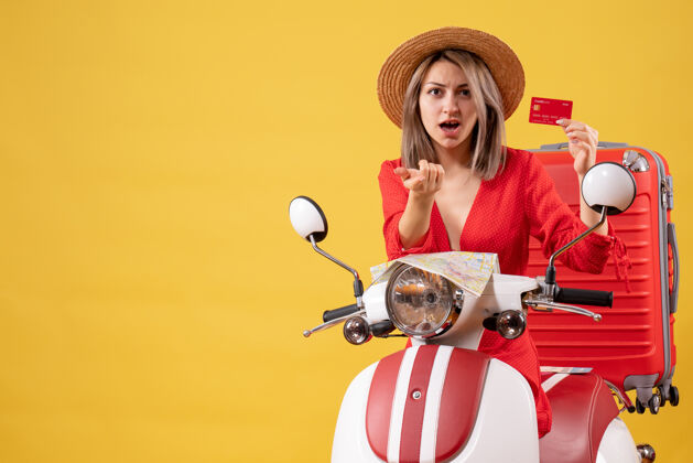 红色前视图穿着红色裙子的年轻女士拿着信用卡指着附近的轻便摩托车行李旅李