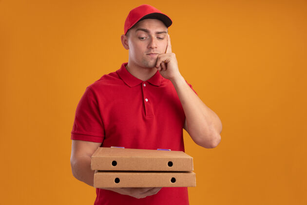 年轻人年轻的送货员穿着制服 戴着帽子 看着隔离在橙色墙上的比萨饼盒感觉衣服脸