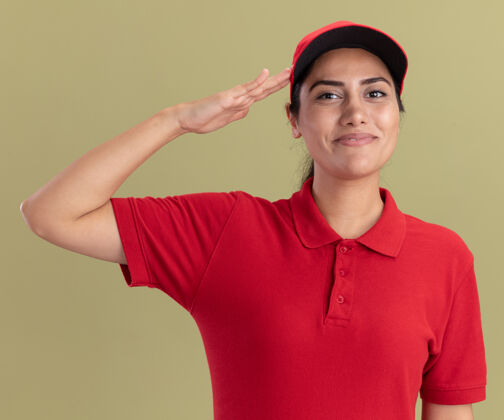 制服身着制服 头戴礼帽 在橄榄绿墙壁上显示敬礼手势的年轻送货女孩穿人感情