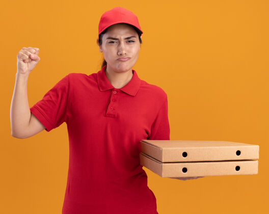 戴着自信的年轻送货女孩穿着制服 戴着帽子 手里拿着披萨盒 举起拳头孤立在橙色的墙上披萨人姿势