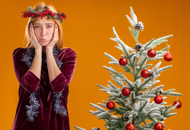 衣服站在圣诞树旁的悲伤的年轻漂亮的女孩穿着红色的裙子 脖子上戴着花环 脸上蒙着花环 手孤立在橙色的墙上站附近花环