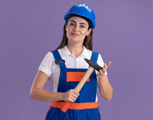 表情身着制服的年轻建筑妇女高兴地把锤子孤立在紫色的墙上人姿势制服