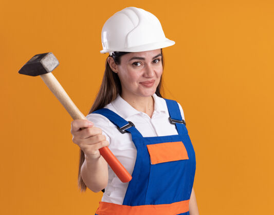 脸身着制服的年轻建筑妇女高兴地向隔离在橙色墙上的摄像机伸出锤子建设者衣服请